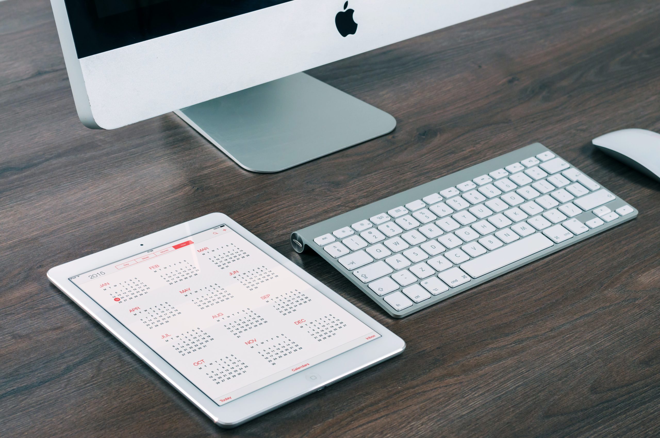 A dark wooden desk. Spread out diagonally are an iPad showcasing a digital calendar, a wireless keyboard and the bottom corner of an iMac